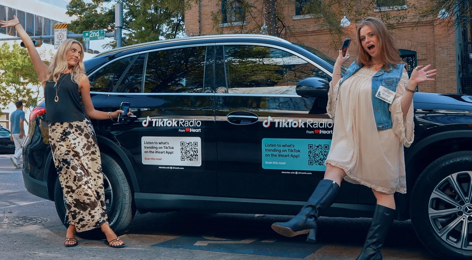 Two people posing beside a black SUV carrying TikTok Radio campaign stickers on a downtown street