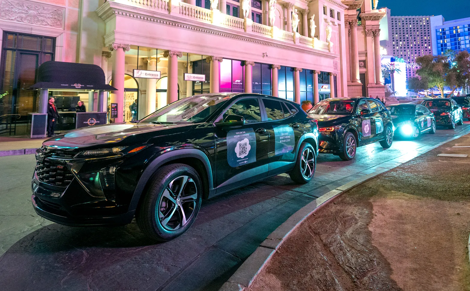 A row of black campaign SUVs with door and hood placements driving along the Las Vegas Strip at night, lit by neon and casino lights